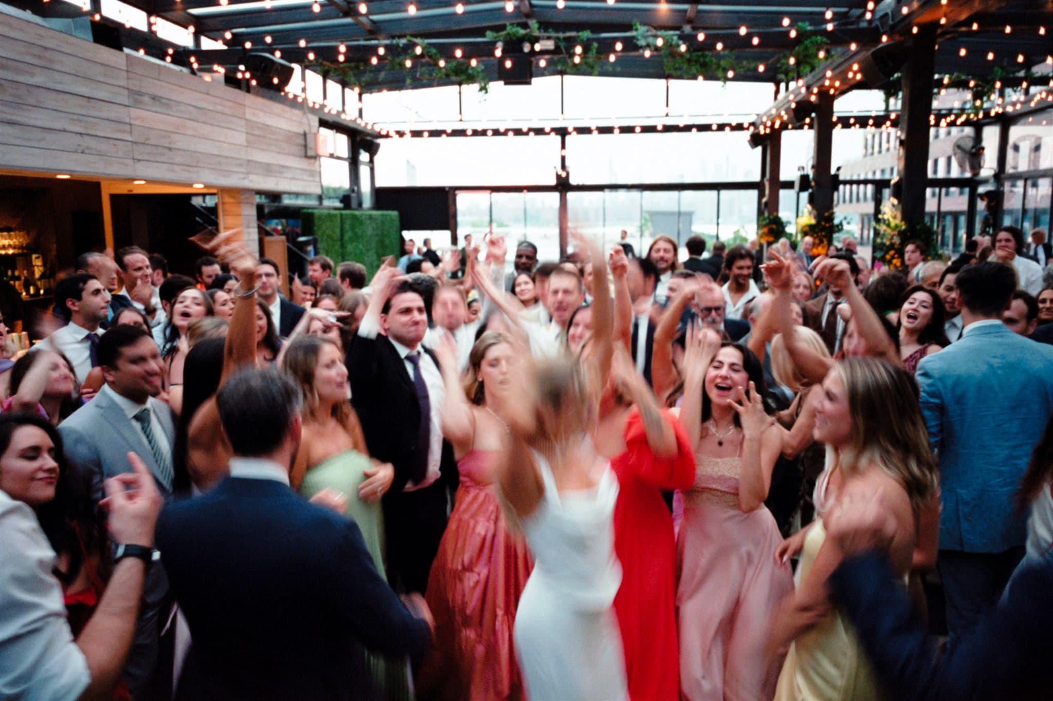 Wedding guests dancing with their hands up on a rooftop under string lights