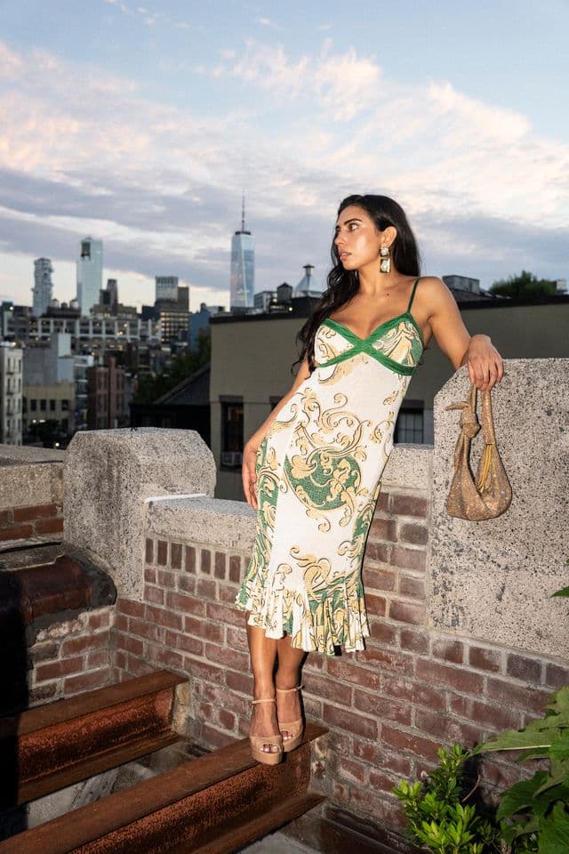 A guest in a green and cream printed dress leaning on a brick rooftop wall above the New York skyline at dusk