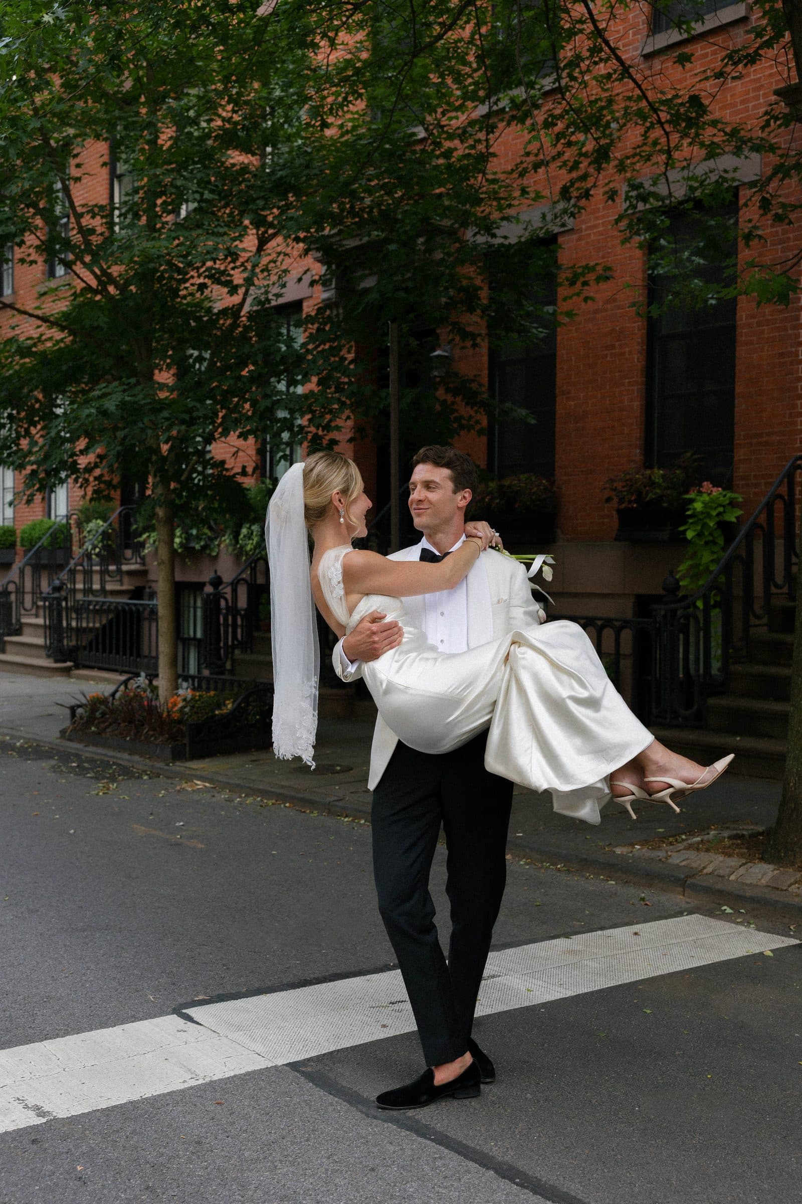 A groom carrying his bride across a tree-lined city street on their wedding day